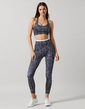 A model in a pair of high-waisted full-length leggings with a hand-sketched safari print, primarily in a dark color, paired with a sports bra and white sneakers.