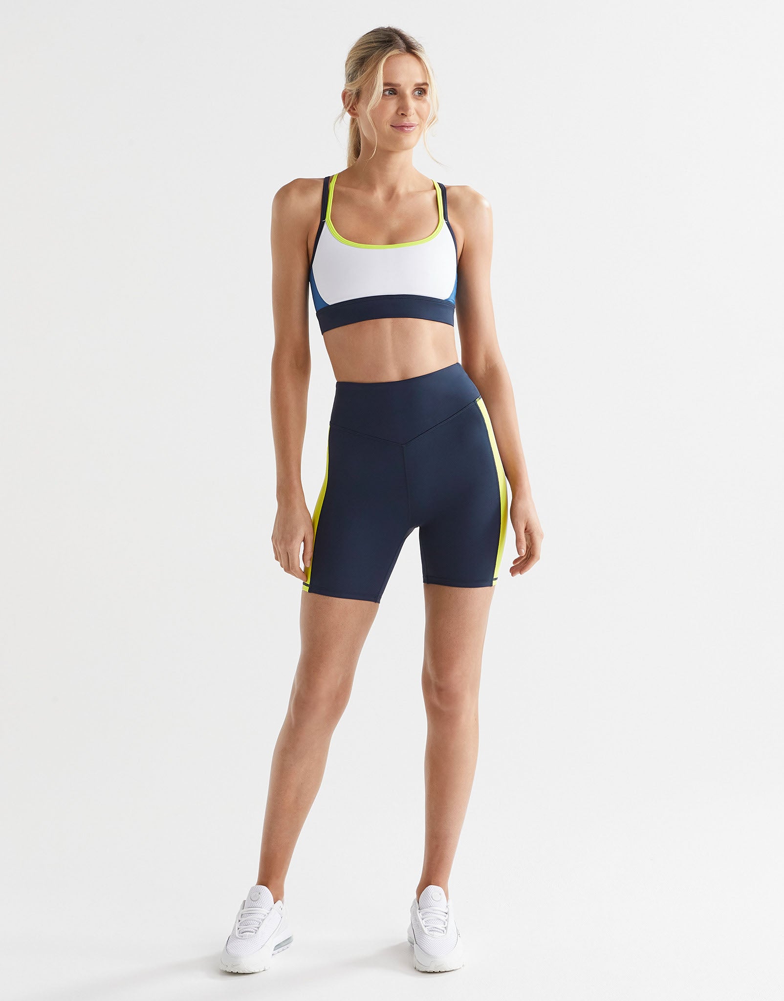A model in a pair of high-waisted navy, lime, and white bike shorts with a reflective silver logo on the back, paired with white sneakers.