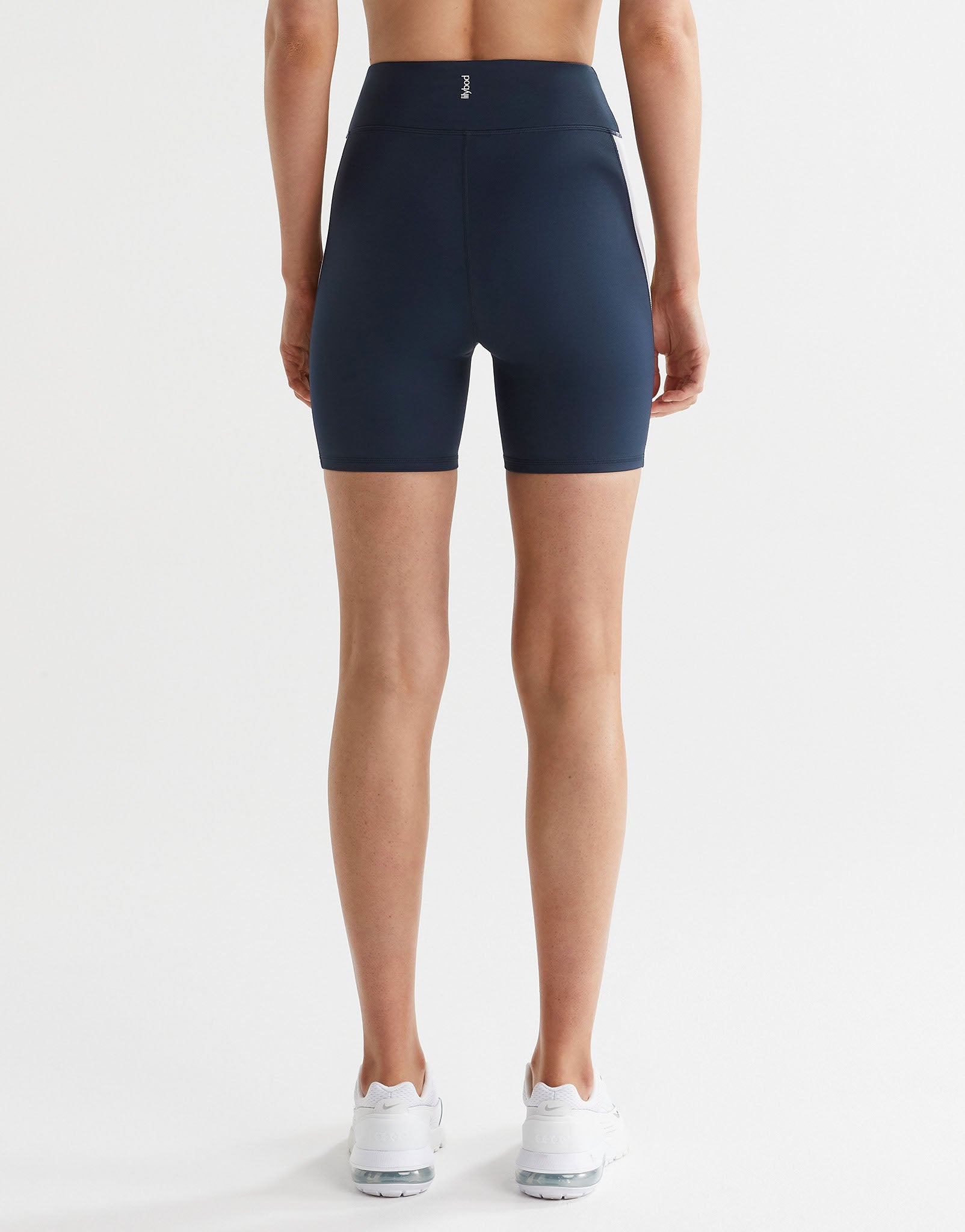 A woman in pair of high-waisted navy, lime, and white bike shorts with a reflective silver logo on the back, paired with white sneakers.