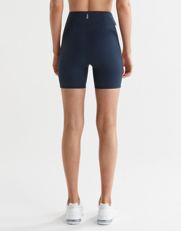 A woman in pair of high-waisted navy, lime, and white bike shorts with a reflective silver logo on the back, paired with white sneakers.