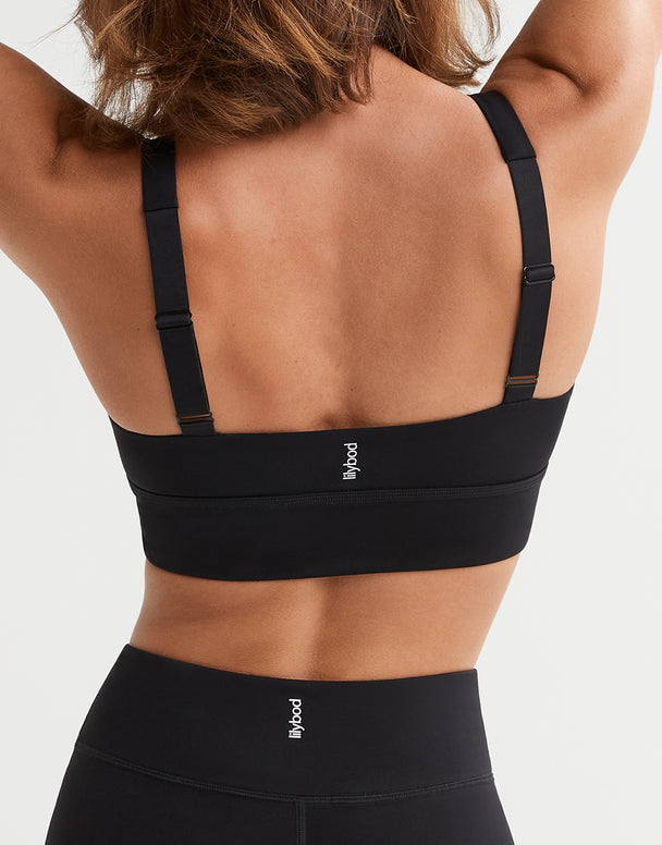 Rear view of a woman wearing a black sports bra and matching high-waisted leggings against a light gray studio background. The brand name 'flywood' is printed in white on the waistband and center back of the bra.