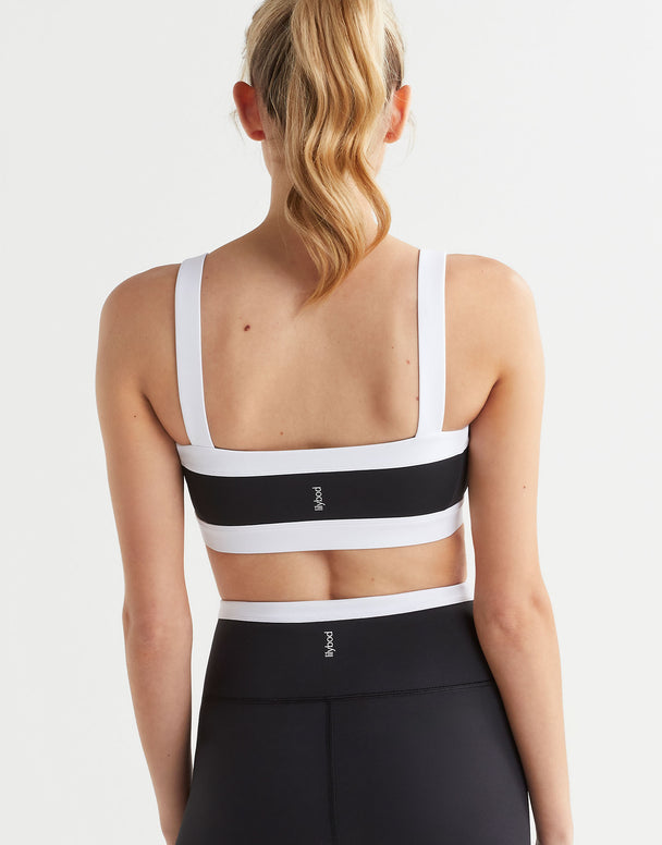 A woman wearing a black and white sports bra with a bandeau style silhouette and the Lilybod logo at the center back and leggings.