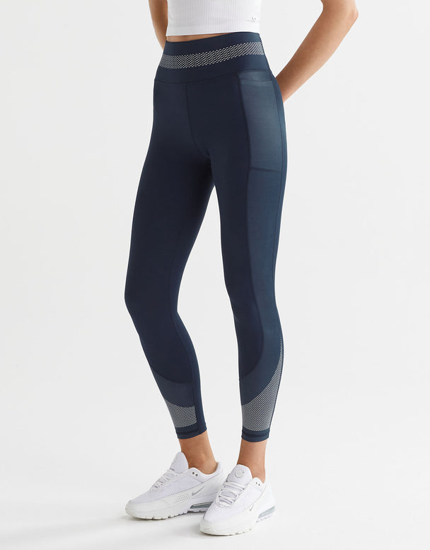 A model wearing navy and white high-waisted leggings with a color-blocked waistband and a printed accent pattern on the legs. She is also wearing a white sports bra and white sneakers.