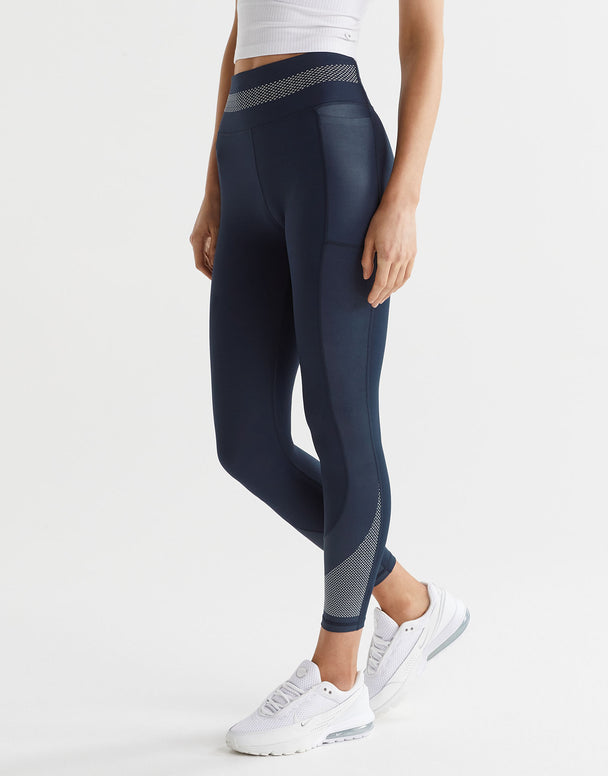 A model wearing navy and white high-waisted leggings with a micro dot waistband and a printed accent pattern on the legs. She is also wearing a white sports bra and white sneakers.