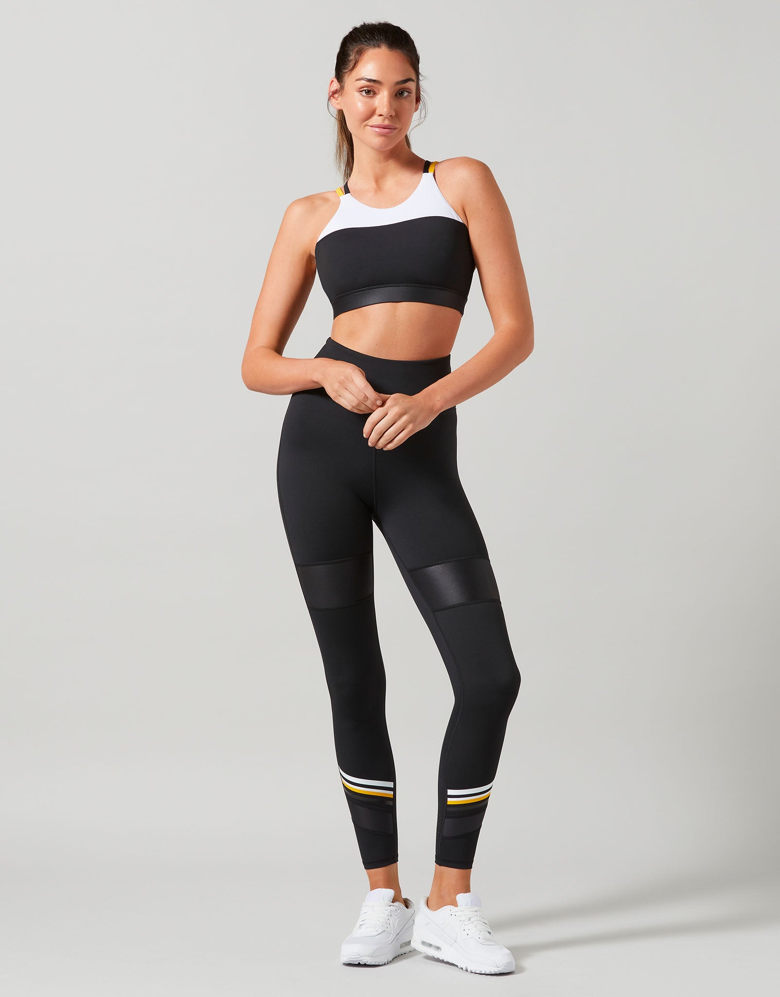 Woman wearing a black sports bra and leggings with white sneakers on a gray background