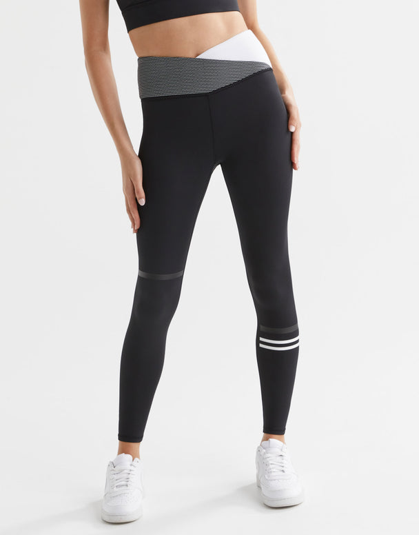 A pair of high-waisted full-length black leggings with a sporty cross-over waistband and a signature Lilybod stripe detail on one leg, paired with a white sports bra and white sneakers.