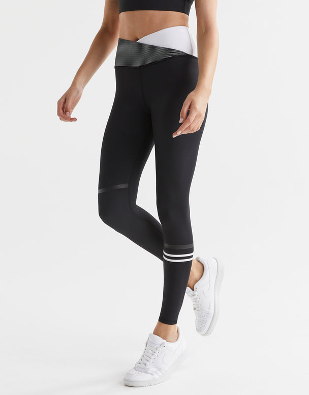A woman wearing a pair of high-waisted full-length black leggings with a sporty cross-over waistband and a signature Lilybod stripe detail on one leg, paired with a white sports bra and white sneakers.