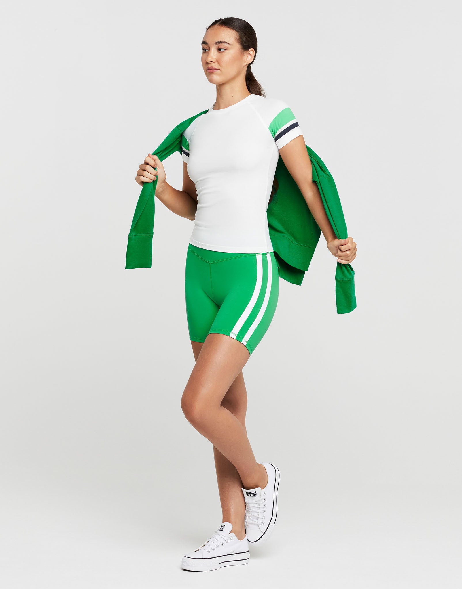 A woman wearing a white and green athletic outfit and white runners on a grey background.