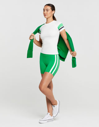 A woman wearing a white and green athletic outfit and white runners on a grey background.