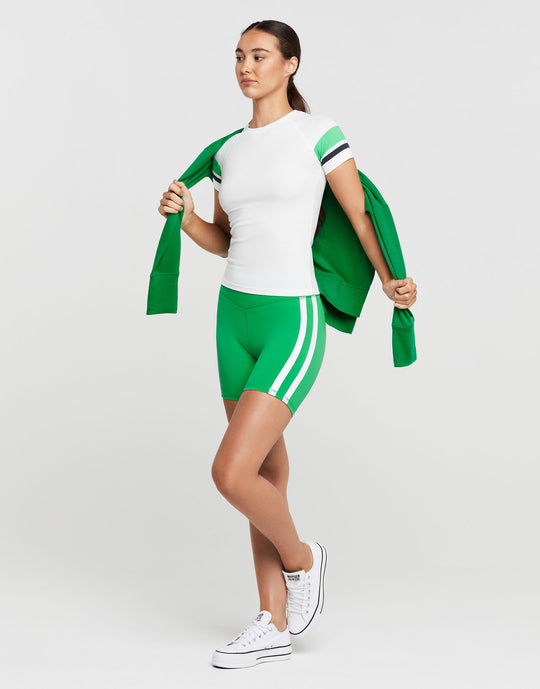 A woman wearing a white and green athletic outfit and white runners on a grey background.