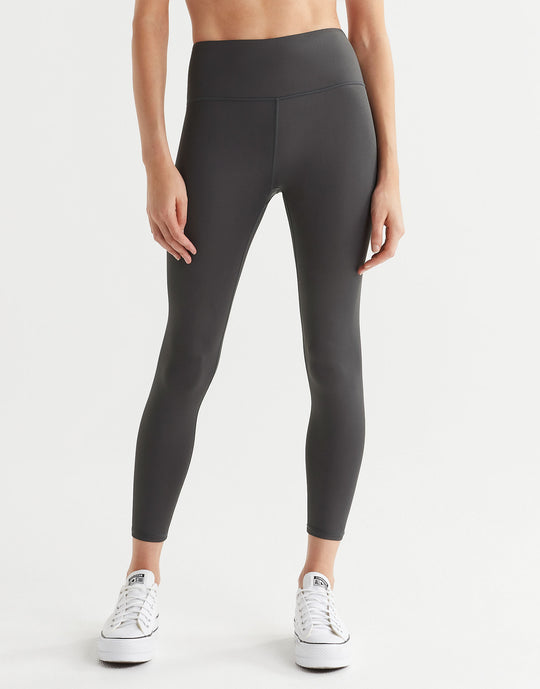 A pair of high shine gunmetal colored leggings, featuring minimal seaming and a logo at the center back. The leggings are presented in a standing position with the wearer's feet facing forward.