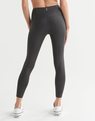 A model wearing a pair of high shine gunmetal colored leggings, featuring minimal seaming and a logo at the center back. 