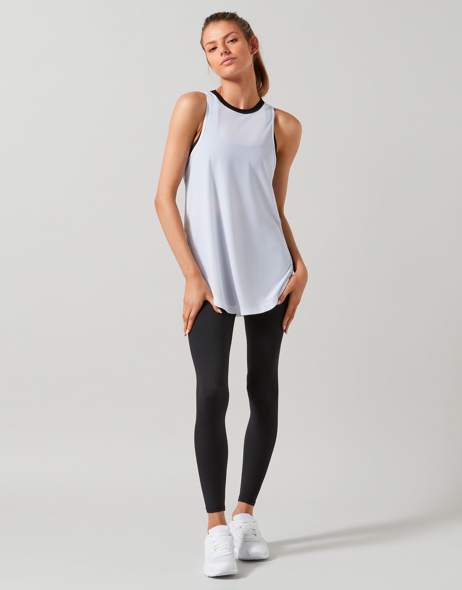 A woman modeling a sleeveless athletic tank top in white with a black crew neck trim, paired with black leggings and white sneakers, against a light gray studio background.