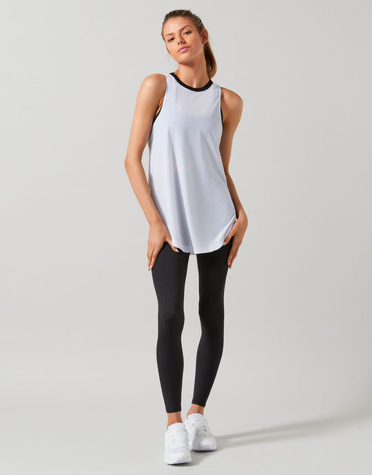 A woman modeling a sleeveless athletic tank top in white with a black crew neck trim, paired with black leggings and white sneakers, against a light gray studio background.