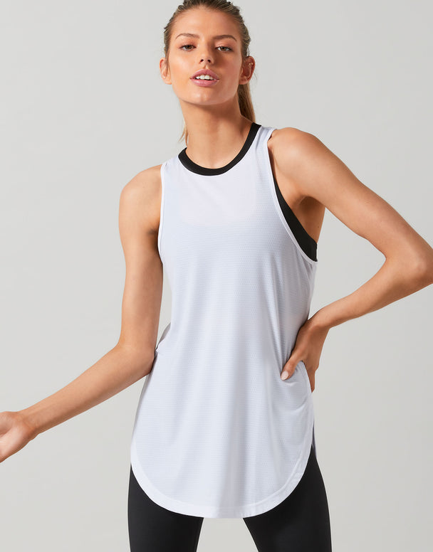 A woman modeling a white sleeveless athletic tank top with black trim at the crew neck and armholes, paired with black leggings, against a light gray studio background.