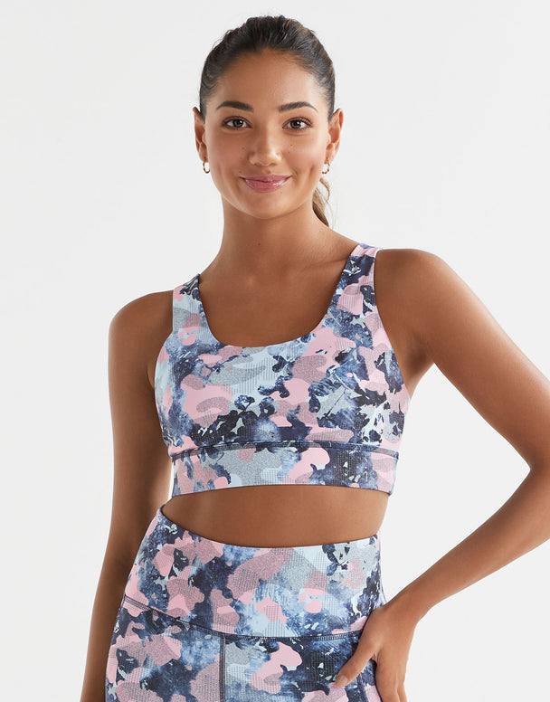 A woman wearing a blue and pink abstract print sports bra with a scoop neck and wide adjustable straps.